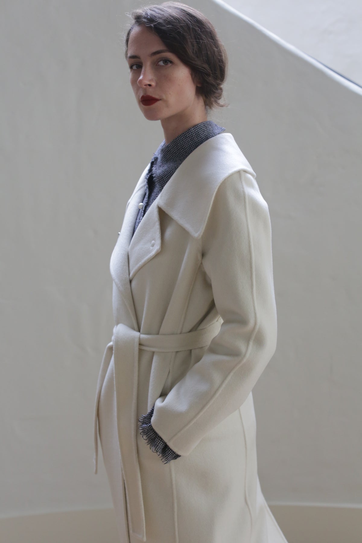 A woman with dark hair and red lipstick is wearing a long, white Malvern Doubleface Cashmere - Undyed wrap coat over a blue collared shirt. The fully lined cashmere coat highlights her serious expression as she stands against a light, neutral background, looking directly at the camera.