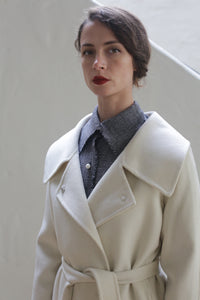 A person with brown hair styled in an updo, wearing a Malvern Doubleface Cashmere - Undyed coat with a wide collar over a checkered shirt. The background is a plain, light-colored wall with part of a staircase visible. The person is looking directly at the camera with a neutral expression.