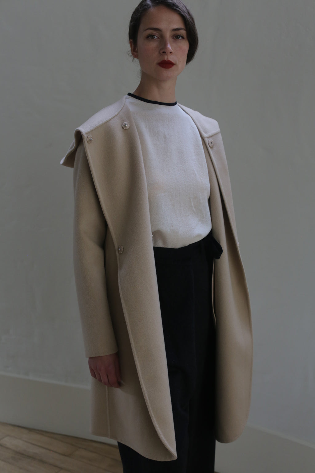 A person stands indoors against a plain wall, wearing a white shirt and beige Mitchley Long Double-face Cashmere | Winter White draped over their shoulders. Their black high-waisted pants complement the ensemble. They have dark hair styled in a low updo and are looking directly at the camera with a neutral expression.