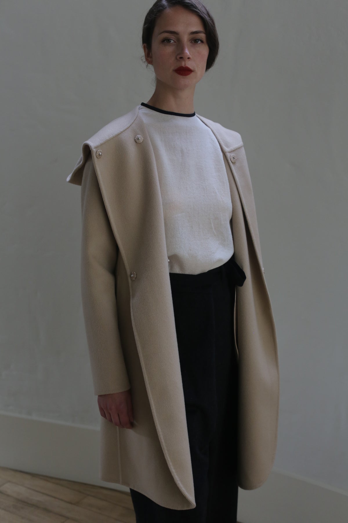 A person stands indoors against a plain wall, wearing a white shirt and beige Mitchley Long Double-face Cashmere | Winter White draped over their shoulders. Their black high-waisted pants complement the ensemble. They have dark hair styled in a low updo and are looking directly at the camera with a neutral expression.