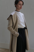 A person stands indoors against a plain backdrop, wearing a Mitchley Long Double-face Cashmere | Winter White over a white top and dark pants. Their hands are in their pockets, and they have a calm expression with eyes closed and lips painted red. The lighting is soft, highlighting the handmade snap buttons on the coat.