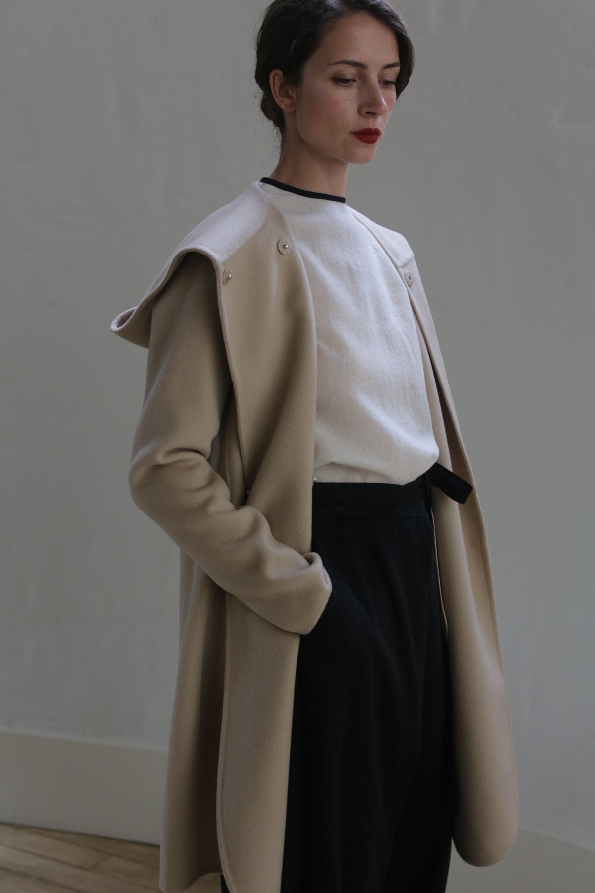 A person stands indoors against a plain backdrop, wearing a Mitchley Long Double-face Cashmere | Winter White over a white top and dark pants. Their hands are in their pockets, and they have a calm expression with eyes closed and lips painted red. The lighting is soft, highlighting the handmade snap buttons on the coat.