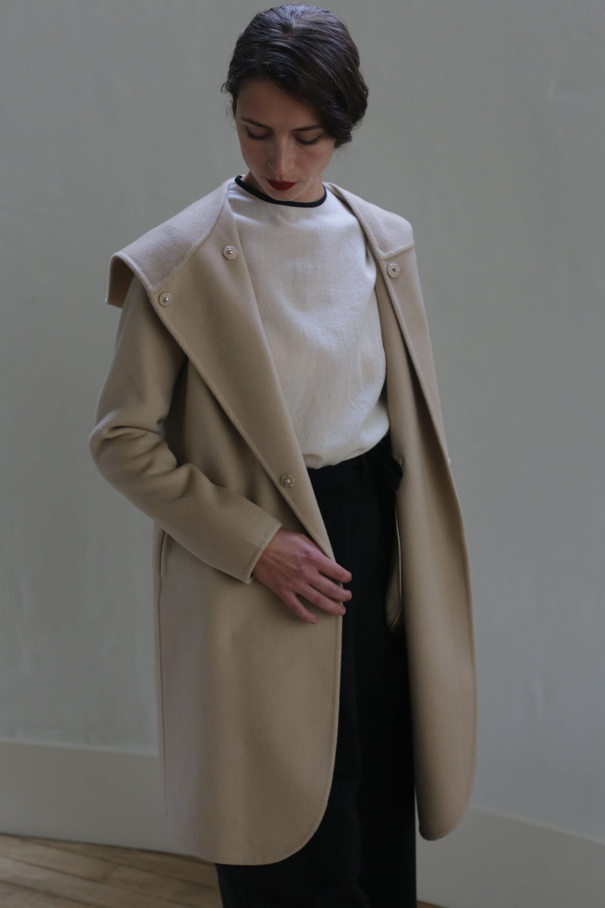 A person with dark hair pulled back is wearing a Mitchley Long Double-face Cashmere | Winter White over a white top and black pants. They are looking downward and have one hand holding the edge of the coat, which features handmade snap buttons. The background is plain and light-colored.