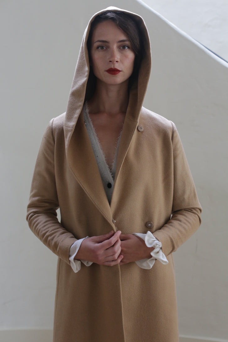 A woman stands facing forward, wearing a Mitchley Long | Camel. The fairytale length coat is buttoned, revealing the white and gray layers underneath. She has red lipstick, a neutral expression, and her hands are positioned near the handmade snap buttons. The background is plain and light-colored.