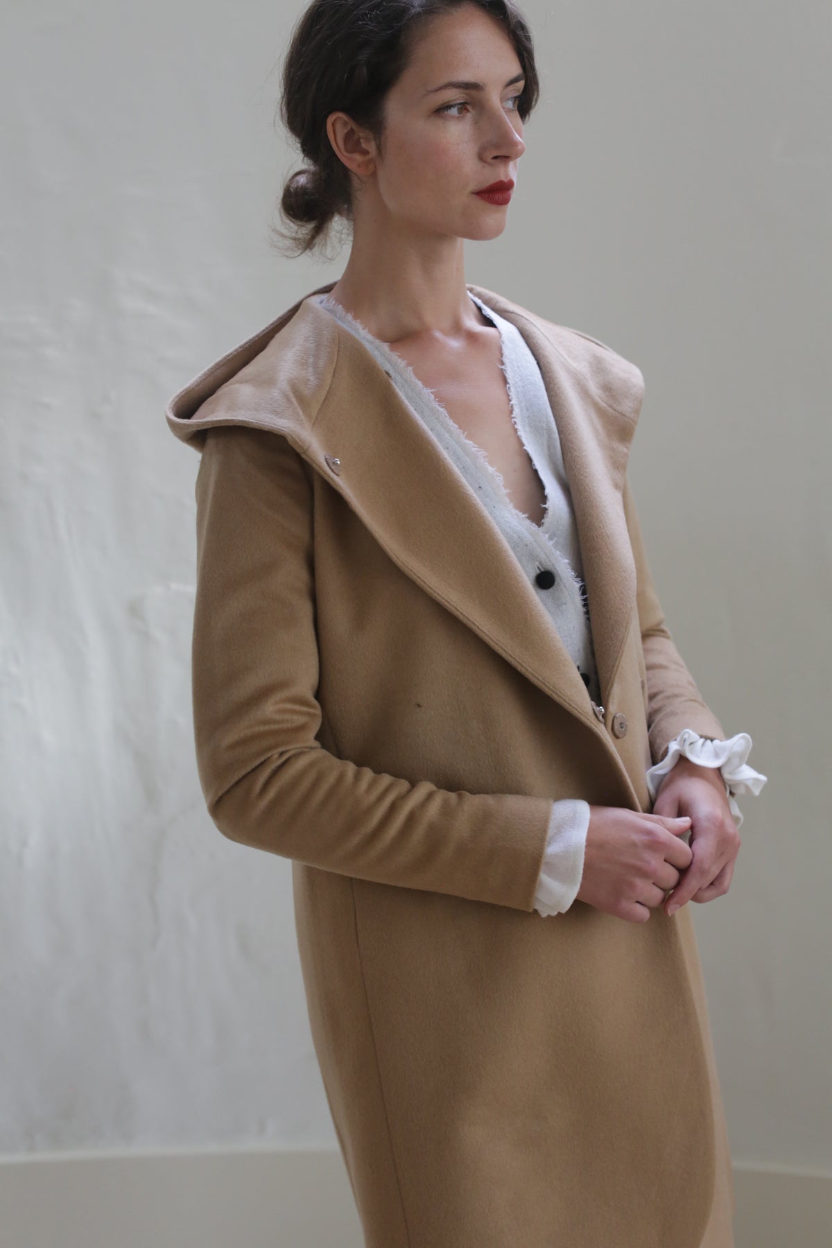 A woman stands against a neutral background, wearing the Mitchley Long | Camel with a hood and handmade snap buttons. She has dark hair pulled back and red lipstick. Underneath the jacket, she wears a white shirt with ruffled cuffs. Her expression is calm and contemplative.