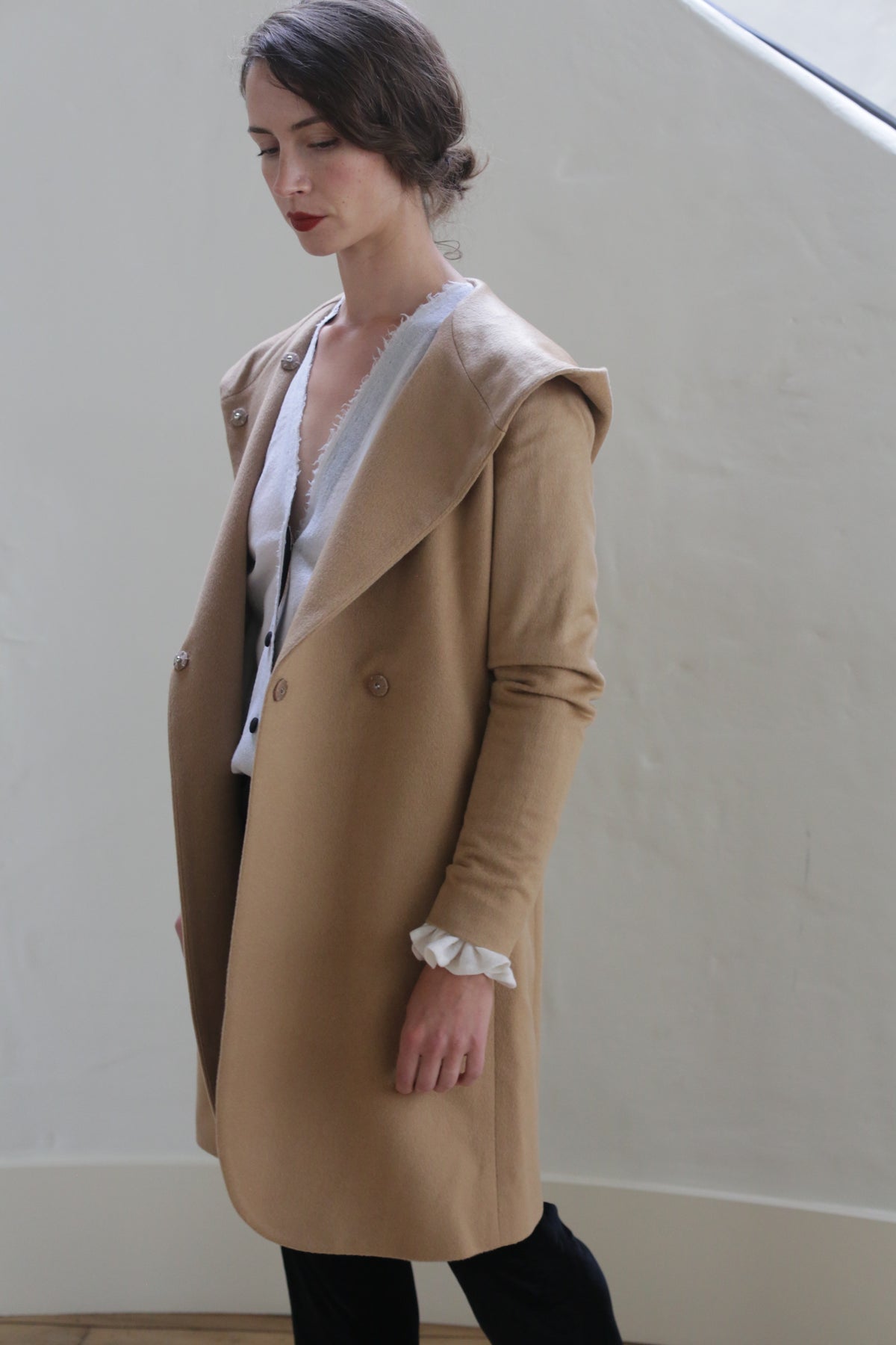 A person stands indoors next to a white wall, wearing a Mitchley Long | Camel with a hood. The person has dark hair pulled back and is looking down with a calm expression. They are wearing a white blouse underneath the coat, which features handmade snap buttons.