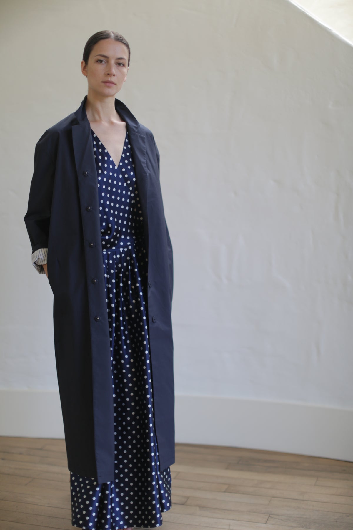 A woman stands indoors with her hands in her pockets, wearing the Painter’s Coat | Navy over a navy and white polka-dot dress made from Typewriter Cotton, against a light-colored wall and wooden floor.
