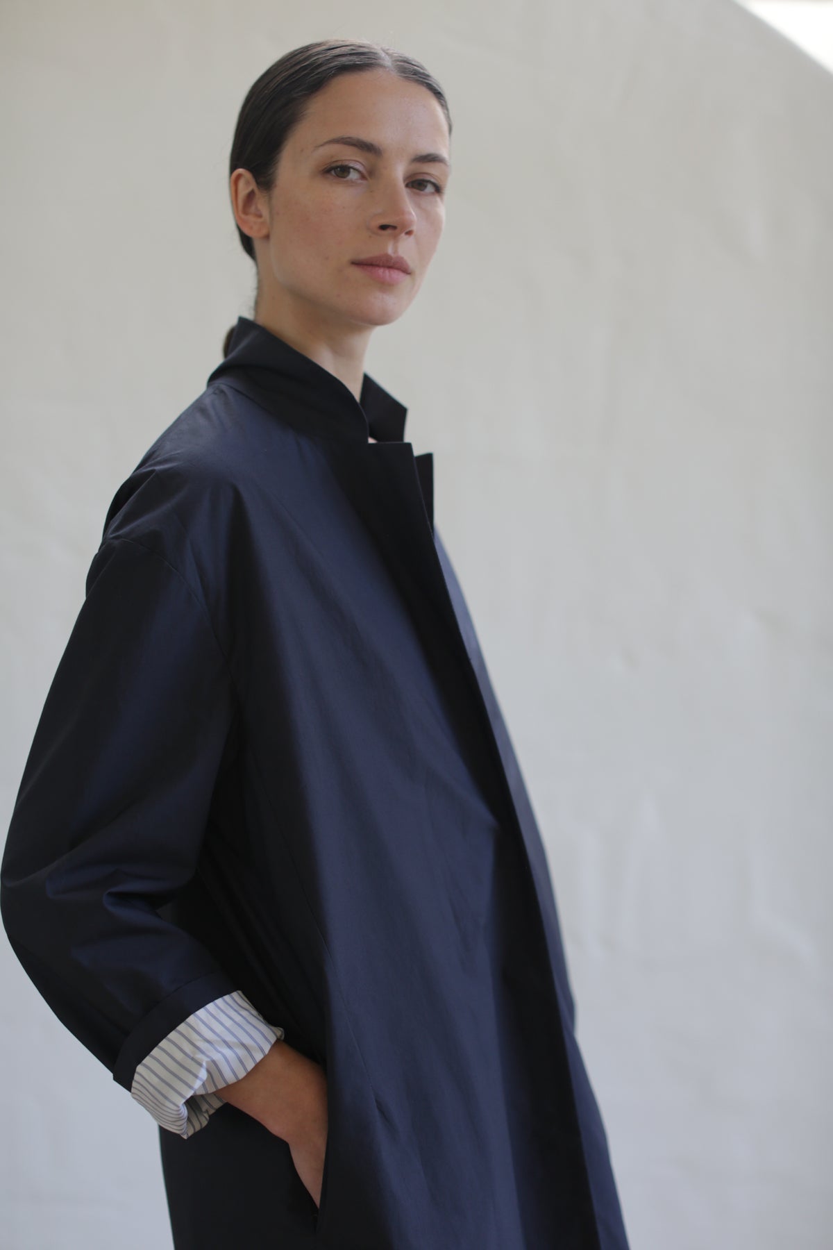A person with dark hair pulled back wears the Painter’s Coat | Navy, its oversized silhouette draping elegantly as they stand with hands in pockets against a light, textured background, gazing confidently at the camera.