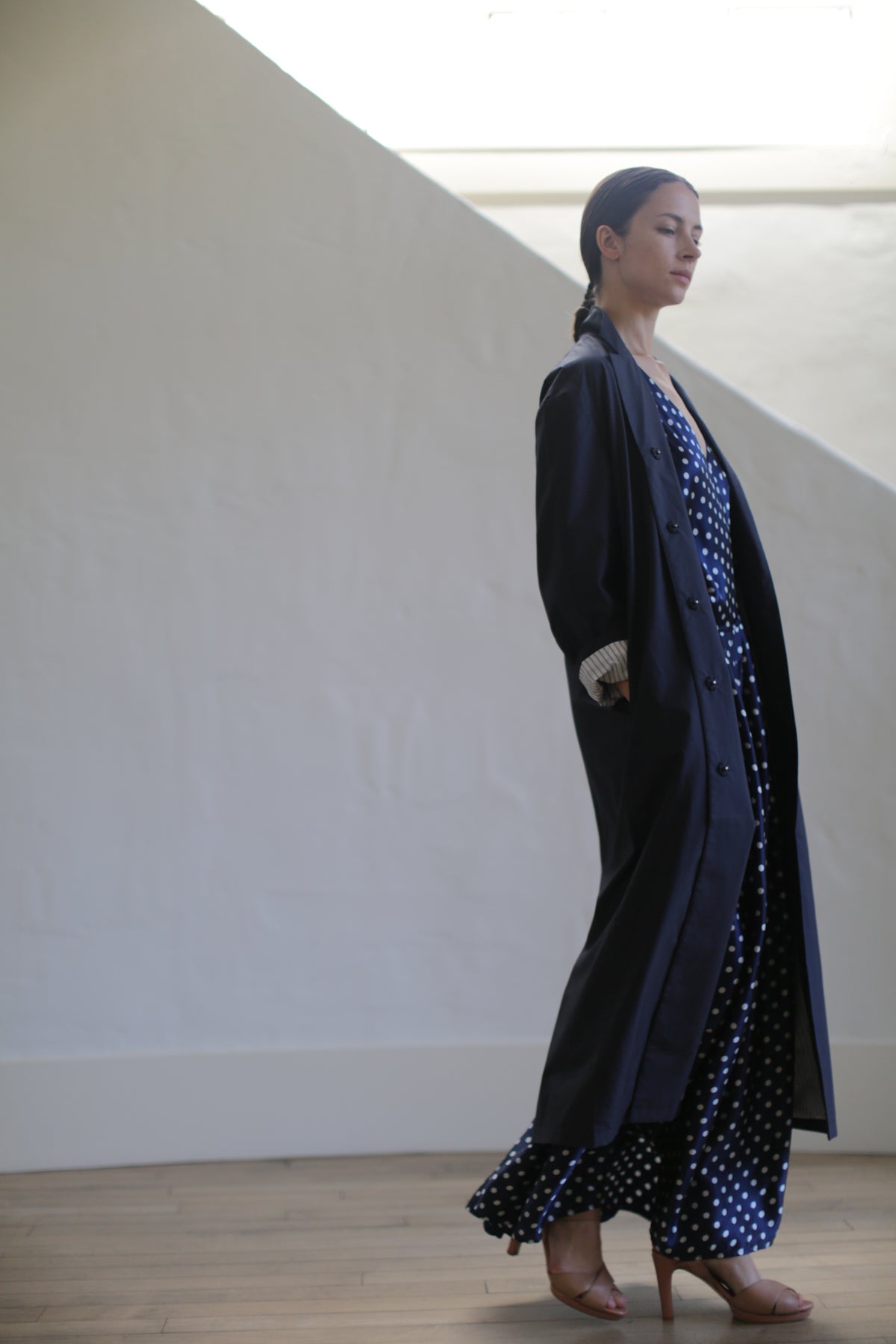 A woman wearing the Painter’s Coat | Navy stands indoors on a wooden floor, eyes closed, with light streaming from above and a simple white background behind her.