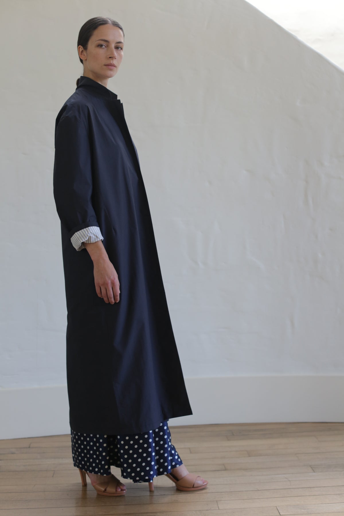 A person stands indoors wearing the Painter’s Coat in Navy with rolled-up sleeves over polka dot pants and tan heels, looking toward the camera against a light-colored wall and wooden floor.
