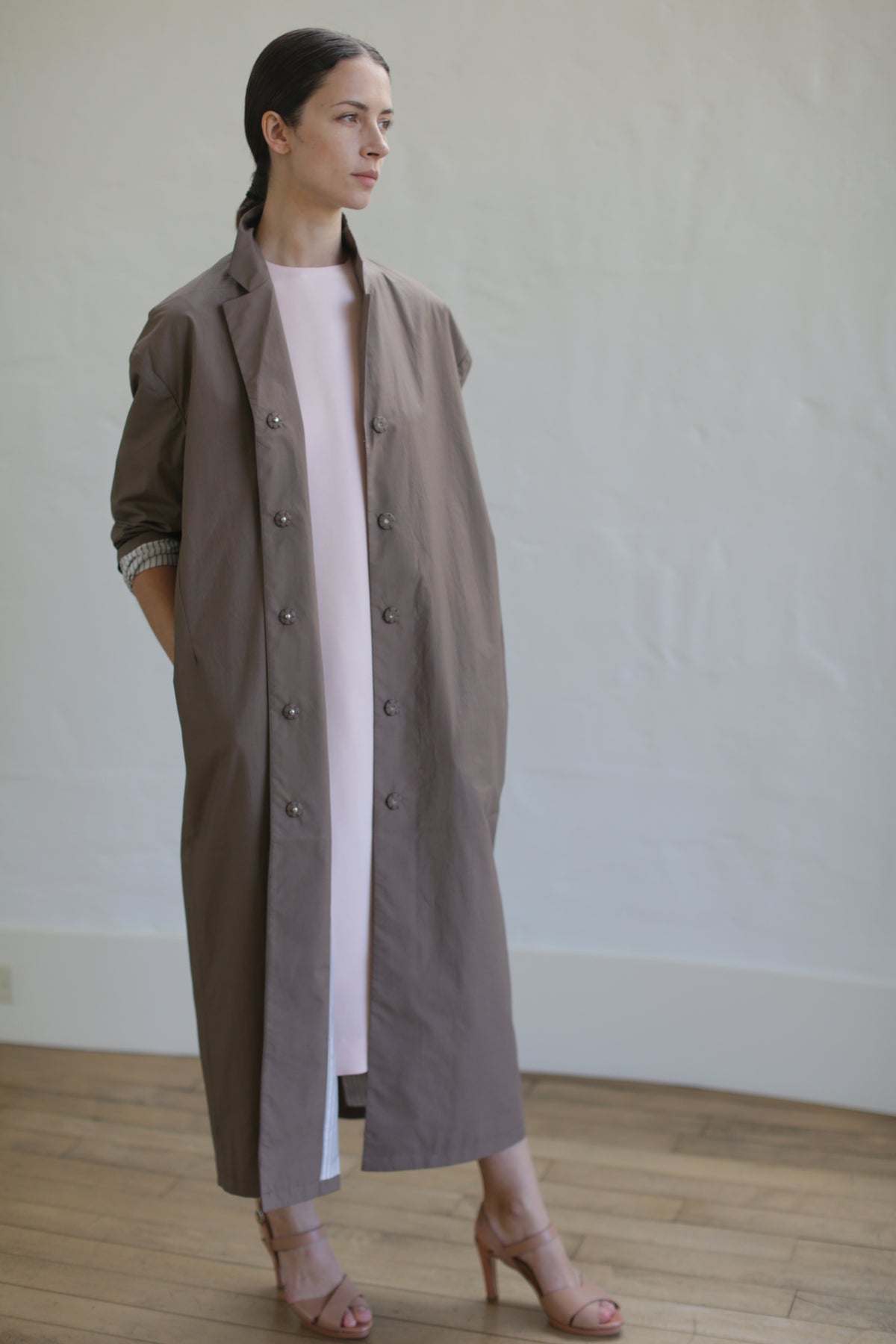 A person stands indoors wearing a Painter's Coat in Clay over a white dress, cinched elegantly with a leather Obi belt. They have dark hair pulled back and are looking to the side, framed by the plain white wall and wood flooring.
