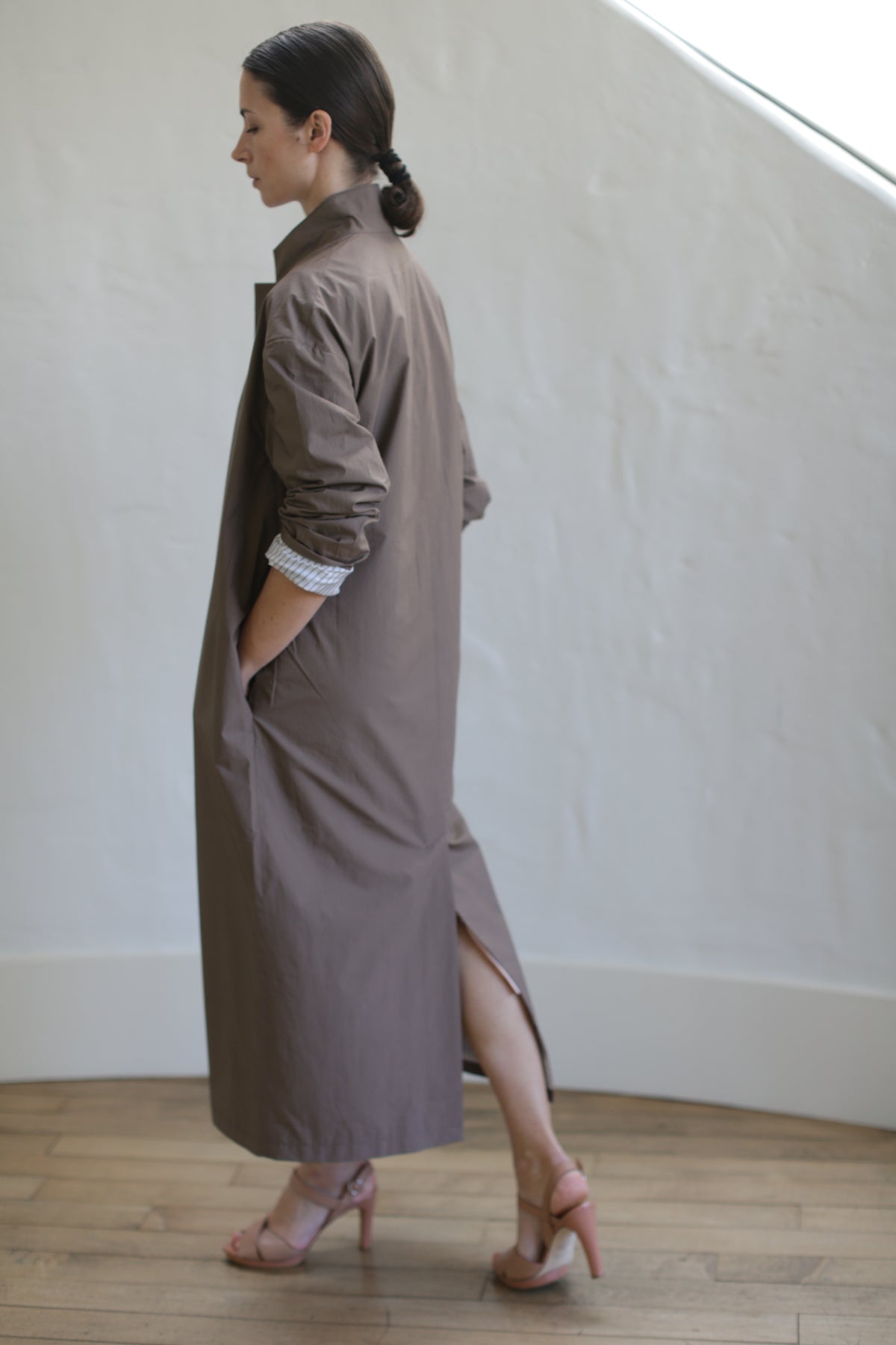 A person stands sideways on a wooden floor, wearing the Painter's Coat in Clay, cinched with a leather Obi belt and paired with pink heels. Their hair is tied back, one hand resting in a pocket. In the background is a white, curved wall softly illuminated by gentle lighting.