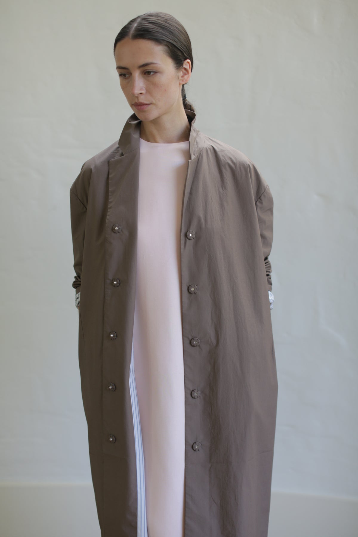 A person stands against a plain background wearing the Painter's Coat in Clay over a light-colored dress. A leather obi belt cinches the waist, adding elegance. Their posture is relaxed, and their dark hair is pulled back neatly.