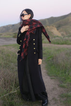 Load image into Gallery viewer, Wearing The Highland Coat—a full-length black coat with gold buttons—a woman stands on a dirt path at dusk, surrounded by green hills and grass. She pairs it with black pants, sunglasses, and a red plaid scarf.