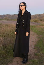 Load image into Gallery viewer, Wearing The Highland Coat, a woman in black sunglasses and boots stands on a dirt path surrounded by grassy hills at sunset. The double-face cashmere outerwear adds elegance to the stunning landscape.