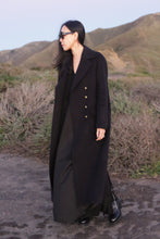 Load image into Gallery viewer, A woman with long dark hair, wearing sunglasses, The Highland Coat in navy double-face cashmere, and black shoes stands outdoors on a path with green hills and shrubs in the background.
