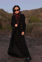 Load image into Gallery viewer, A woman wearing The Highland Coat stands outdoors on a rocky path, paired with black attire and a red plaid scarf, with shrubs and hills in the background.