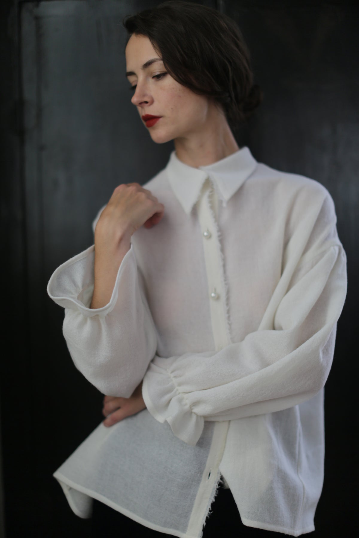 A person with dark hair is standing against a dark background, wearing a loose-fitting, long-sleeved Poet Shirt - Japanese Wool Gauze | Undyed with buttons. They have a thoughtful expression while looking down and resting one hand on their neck. Their red lipstick adds a pop of color.
