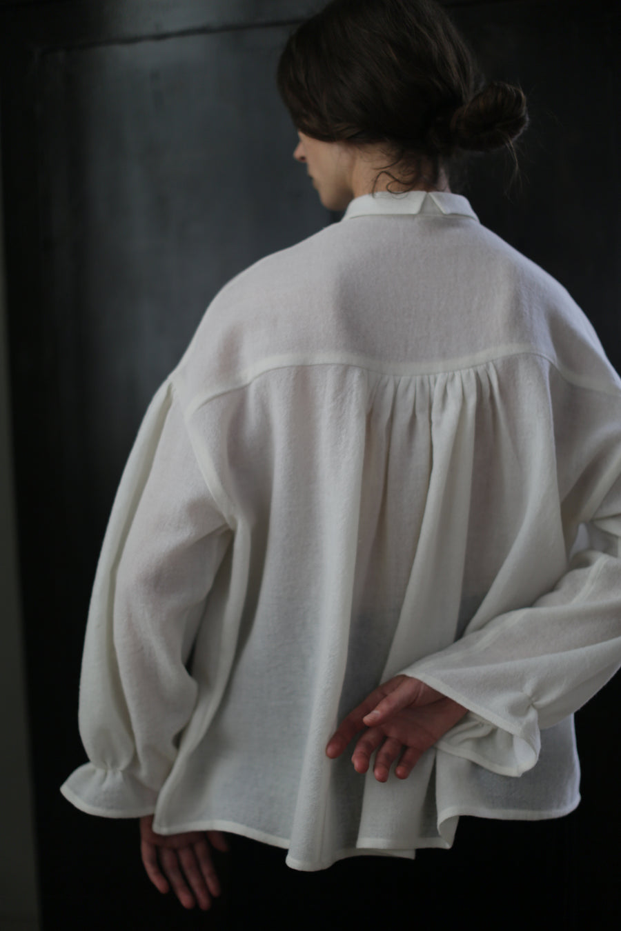 A person with dark hair styled in a bun is seen from the back, wearing a loose-fitting, white Poet Shirt - Japanese Wool Gauze | Undyed. The person has one hand behind their back while standing against a dark, blurred background. The blouse appears to be crafted from Japanese wool gauze, emphasizing sustainable design.
