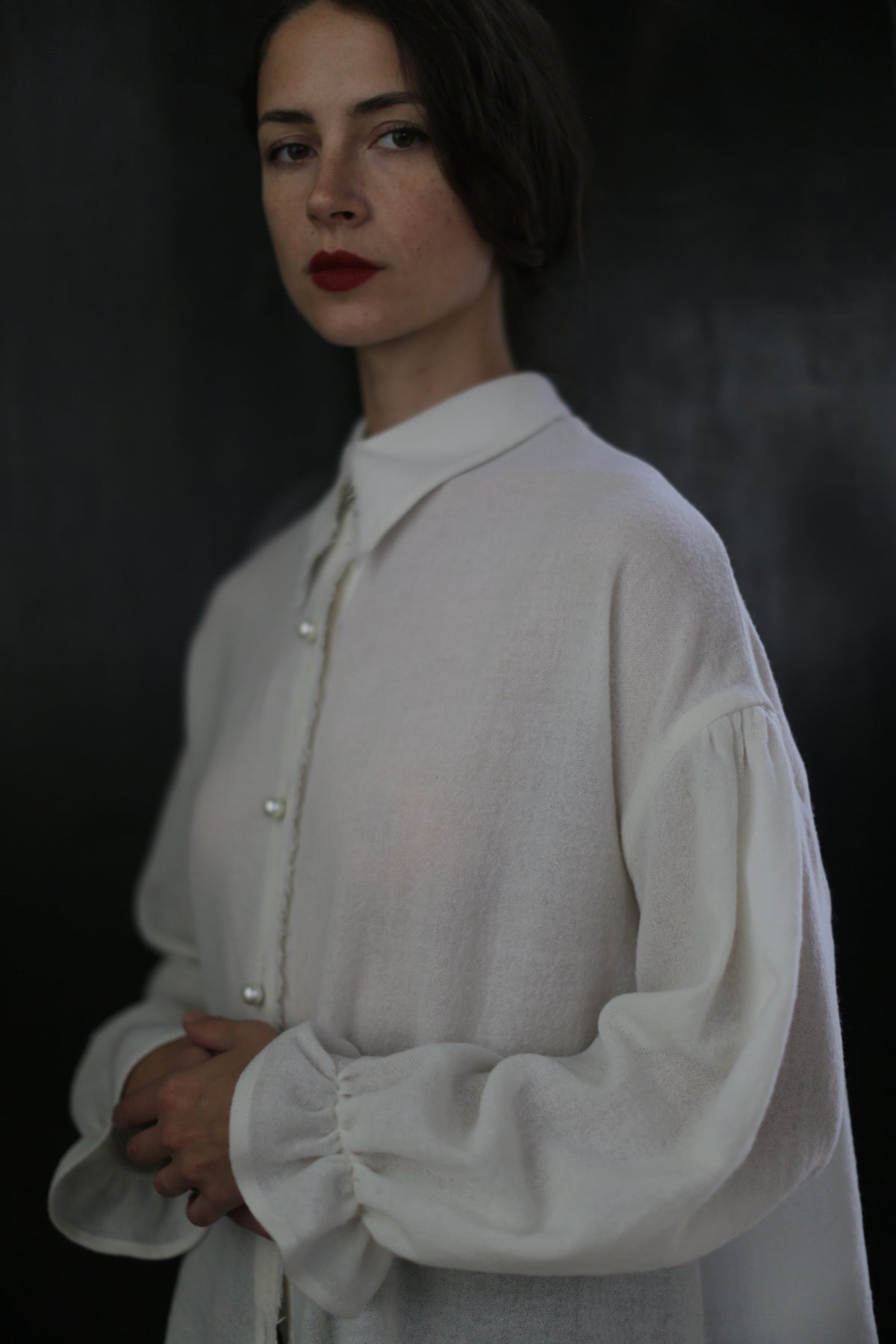 A woman with dark hair stands against a dark background, wearing a loose-fitting Poet Shirt - Japanese Wool Gauze | Undyed. Her blouse has long, puffy sleeves and a high collar. With bold red lipstick and a neutral expression, she embodies the essence of sustainable design.