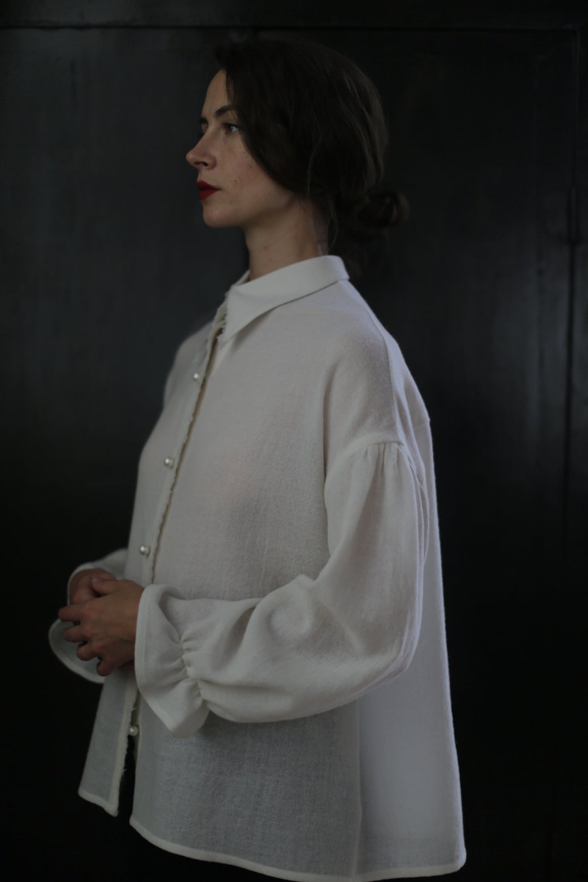 A woman with dark hair tied back, wearing a white Poet Shirt - Japanese Wool Gauze | Undyed with puffy sleeves made from Japanese Wool Gauze, stands in profile against a dark background. She appears contemplative, with her hands loosely clasped in front of her.