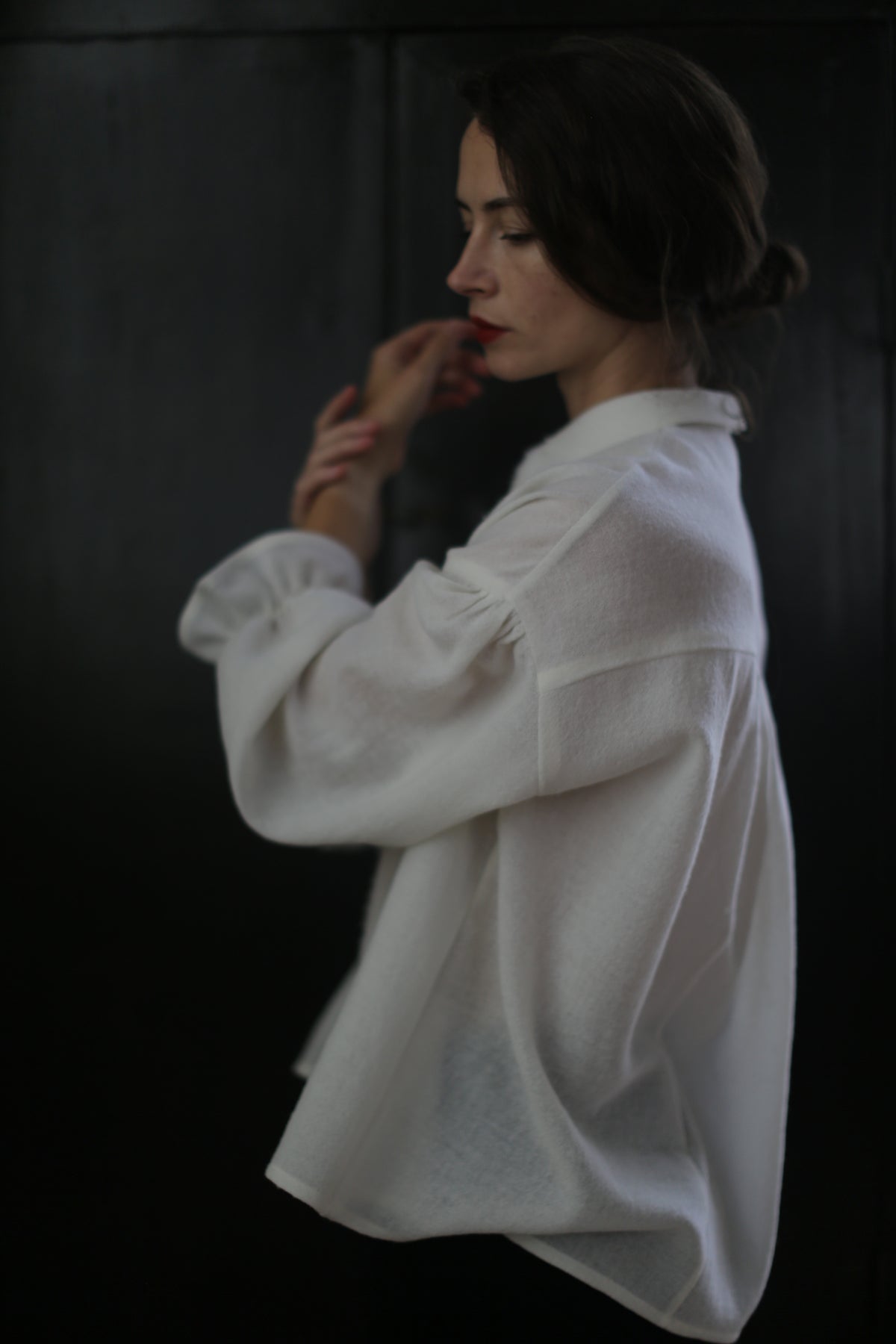 A woman with dark hair is standing sideways against a dark background. She is wearing an oversized Poet Shirt - Japanese Wool Gauze | Undyed, its long sleeves draping elegantly. Her hair is loosely tied back, and she appears to be lost in thought, with her hands gently touching her face.
