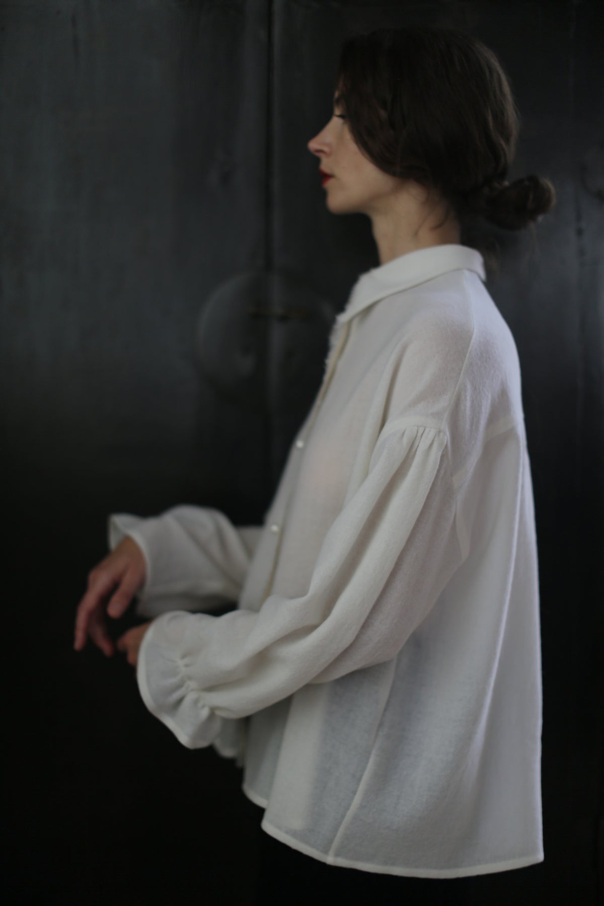 A person with dark hair styled in a low bun is standing in profile against a dark background. They are wearing a light-colored, Poet Shirt - Japanese Wool Gauze | Undyed with loose-fitting, billowy sleeves and a collar, exemplifying sustainable design. Their hands are relaxed and positioned slightly forward.