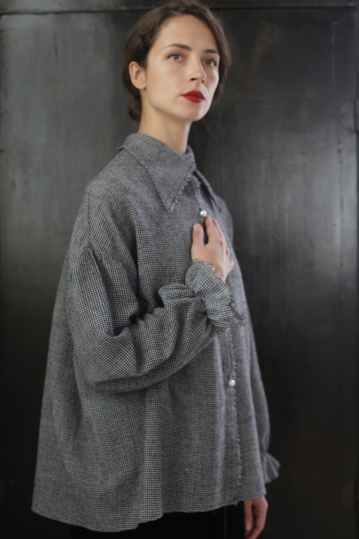 A woman with brown hair stands in front of a dark background, wearing the Poet Shirt - Japanese Wool Gauze | BW Houndstooth. She gazes slightly upward with a serious expression, resting her left hand on her chest, exuding a Byronesque flair.