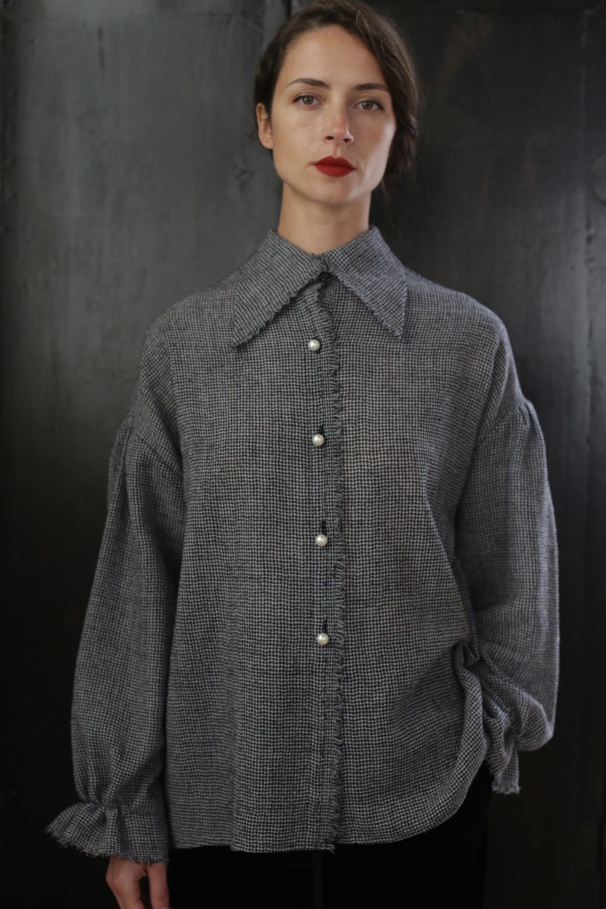 A woman with dark hair tied back stands against a dark background, wearing a textured, oversized Poet Shirt - Japanese Wool Gauze | BW Houndstooth with pearl-like buttons. She has a calm expression and red lipstick, with her left hand partially tucked into her pocket, exuding a Byronesque flair.