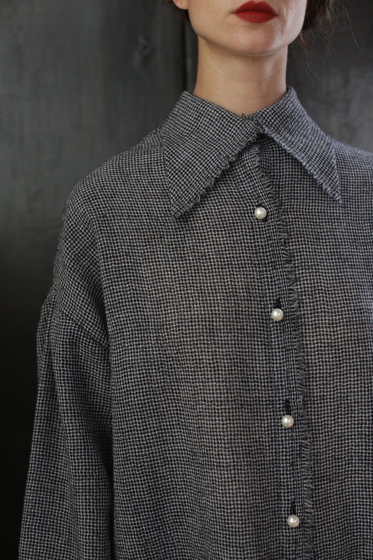 Close-up of a person wearing a Poet Shirt - Japanese Wool Gauze | BW Houndstooth, featuring pearl-like buttons. The background is dark, and the person's face is partially visible, showing red lipstick and a neutral expression, exuding a hint of Byronesque flair.