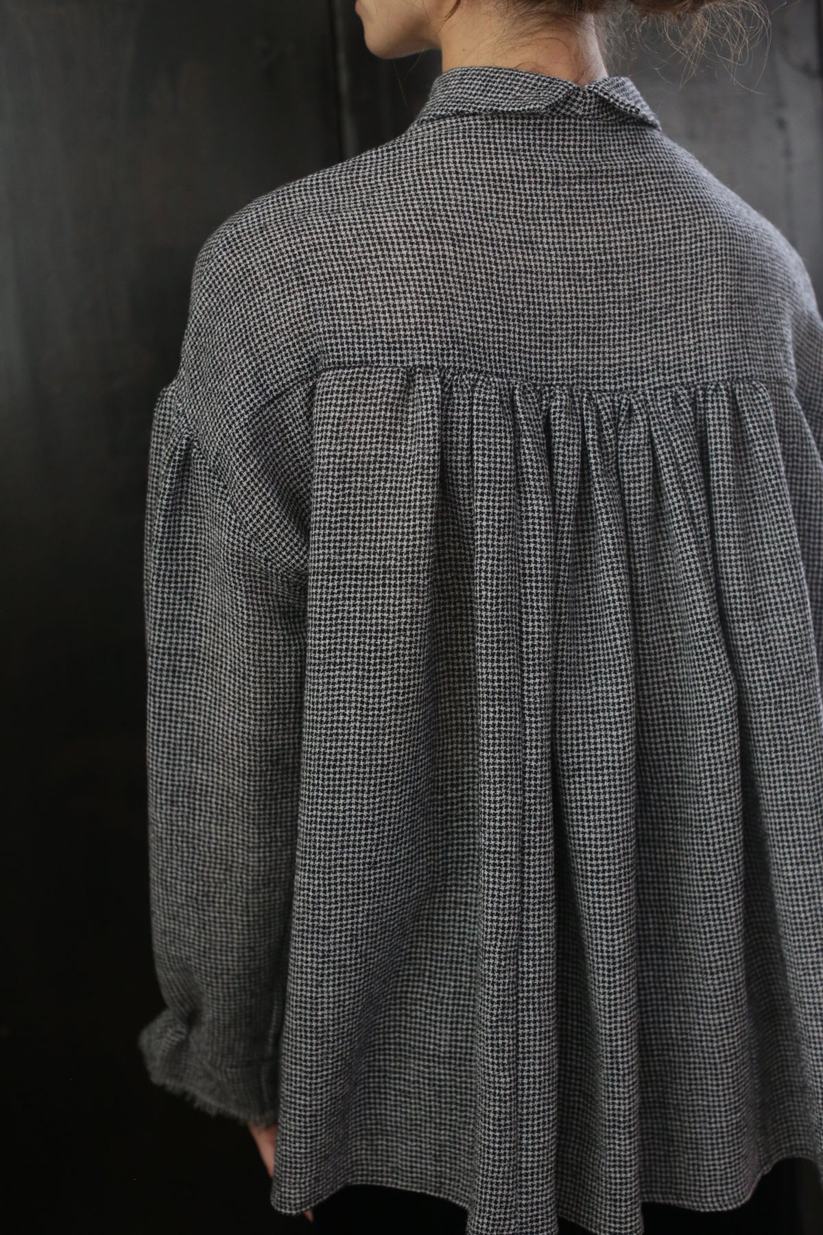 A person stands with their back to the camera, wearing a loose-fitting, black and white checkered Poet Shirt - Japanese Wool Gauze | BW Houndstooth. The blouse has a gathered detail at the back yoke and long sleeves, exuding Byronesque flair. The background is dark and out of focus.