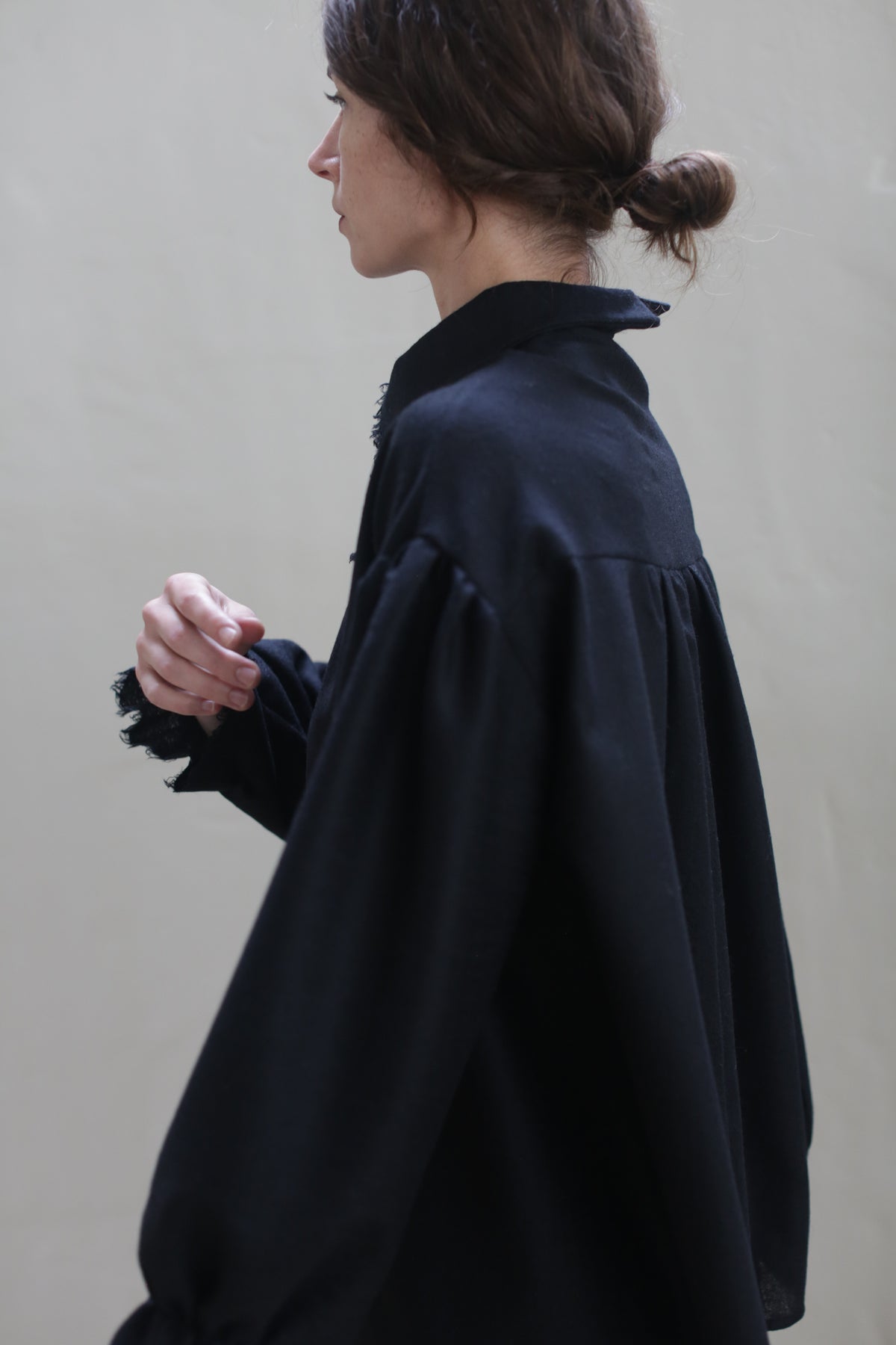 A person with brown hair tied in a low bun is pictured from the side wearing a loose-fitting Poet Shirt - Japanese Wool Gauze | Black with gathered sleeves. The beige background is plain, emphasizing the simplicity and sustainable design of the elegant outfit.