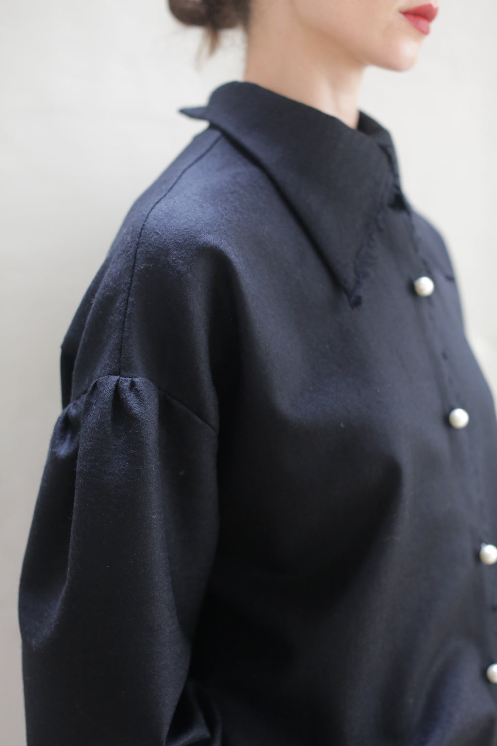A close-up of a person wearing a dark, long-sleeved Poet Shirt - Japanese Wool Gauze | Black with pearl-like buttons. The handmade shirt, crafted from Japanese wool gauze, features a broad collar and slightly puffed sleeves. Their hair is tied back, and only part of their face and upper body is visible against the plain, light background.