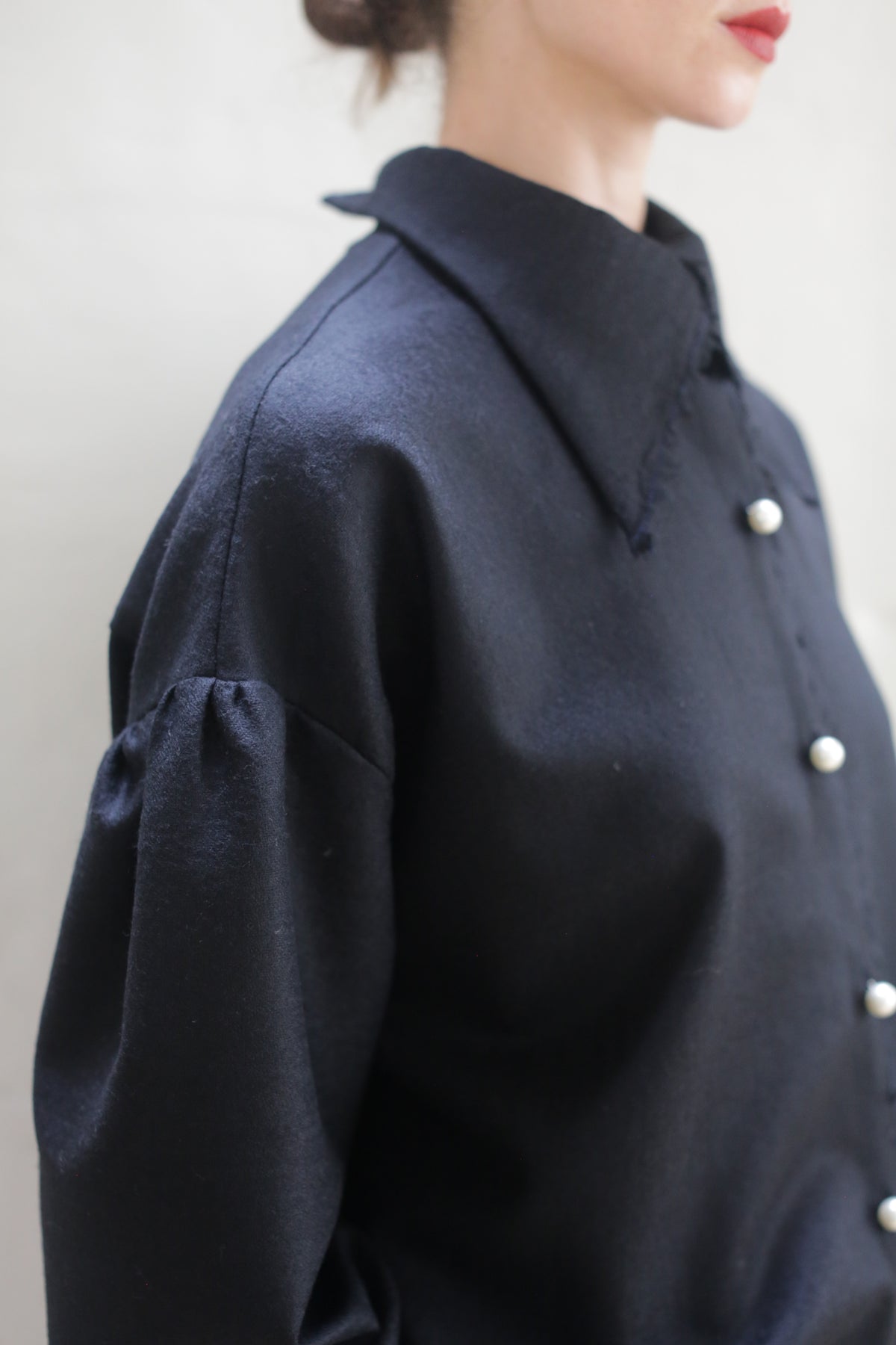 A close-up of a person wearing a dark, long-sleeved Poet Shirt - Japanese Wool Gauze | Black with pearl-like buttons. The handmade shirt, crafted from Japanese wool gauze, features a broad collar and slightly puffed sleeves. Their hair is tied back, and only part of their face and upper body is visible against the plain, light background.