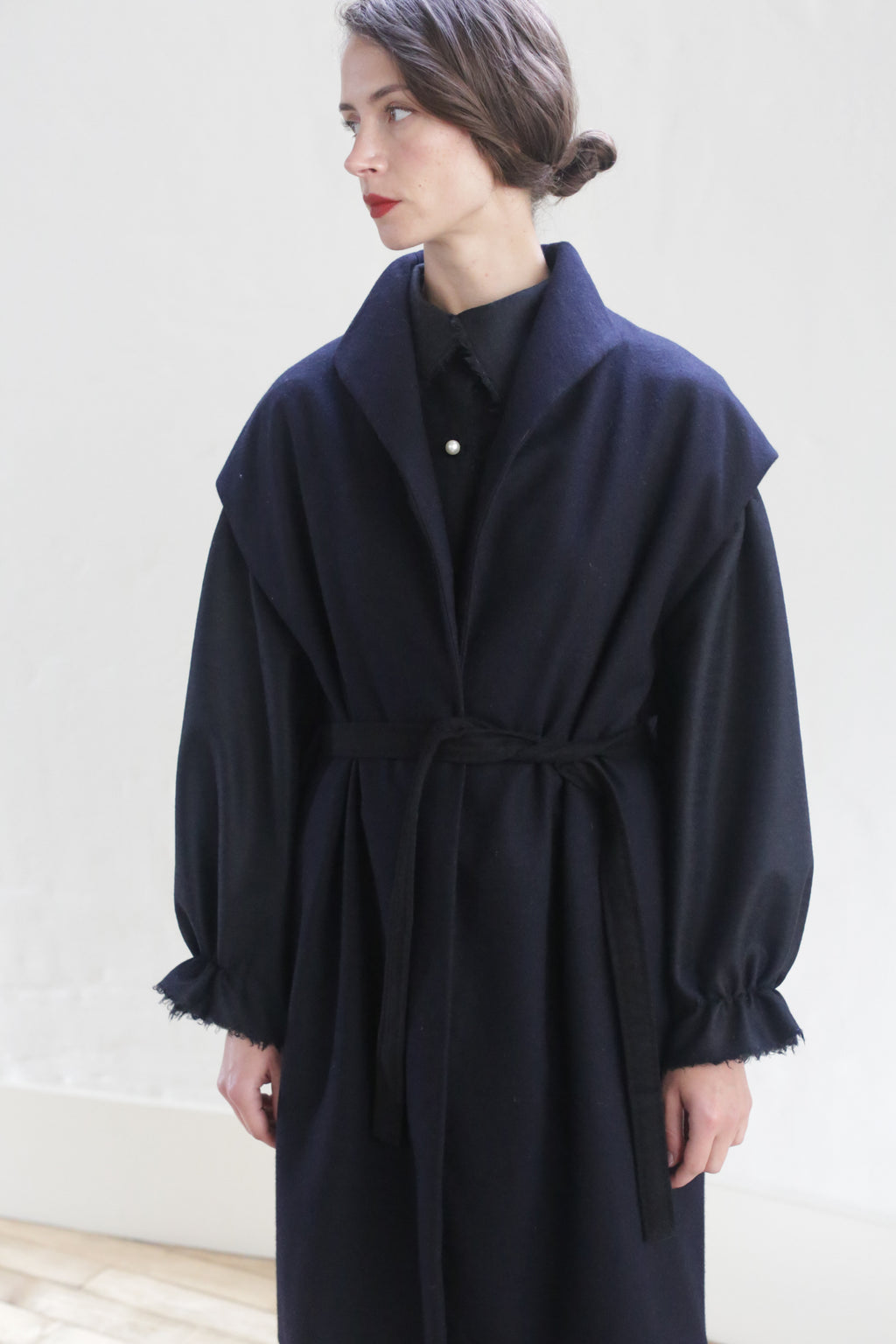 A person with tied-back hair is standing against a white background, wearing a Poet Vest Coat | Navy. This dark, long-sleeved, handmade cashmere coat features a tied waist and ruched cuffs over a button-up shirt. The person is looking to the side with a neutral expression.