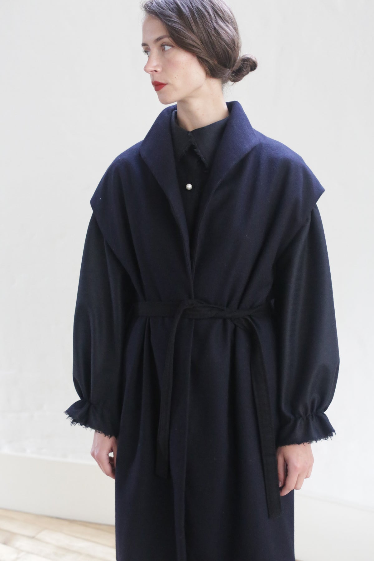 A person with tied-back hair is standing against a white background, wearing a Poet Vest Coat | Navy. This dark, long-sleeved, handmade cashmere coat features a tied waist and ruched cuffs over a button-up shirt. The person is looking to the side with a neutral expression.