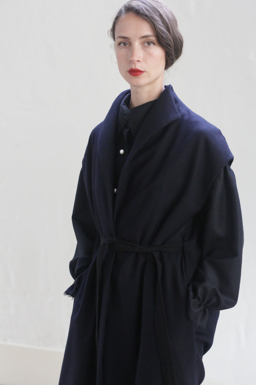A person with a serious expression stands against a plain background. They have dark hair, pulled back, and wear a Poet Vest Coat | Navy with their hands in the pockets. They are also wearing a dark shirt underneath and have bold red lipstick.