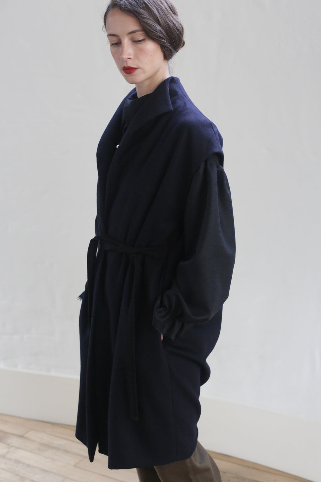 A person with short dark hair, wearing an elegant handmade Poet Vest Coat | Navy by Atelier Lan Jaenicke, stands against a plain light-colored wall. The belt tied at the waist accentuates the coat’s design. The person appears to be looking downwards with a thoughtful expression. The floor is wooden.