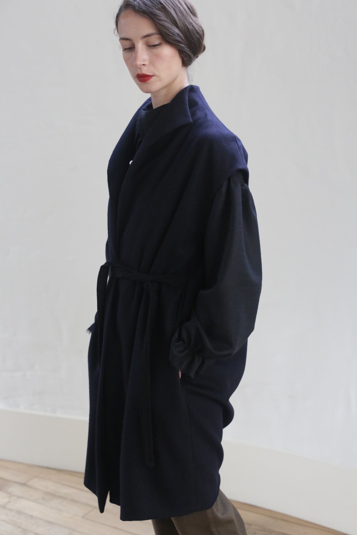 A person with short dark hair, wearing an elegant handmade Poet Vest Coat | Navy by Atelier Lan Jaenicke, stands against a plain light-colored wall. The belt tied at the waist accentuates the coat’s design. The person appears to be looking downwards with a thoughtful expression. The floor is wooden.