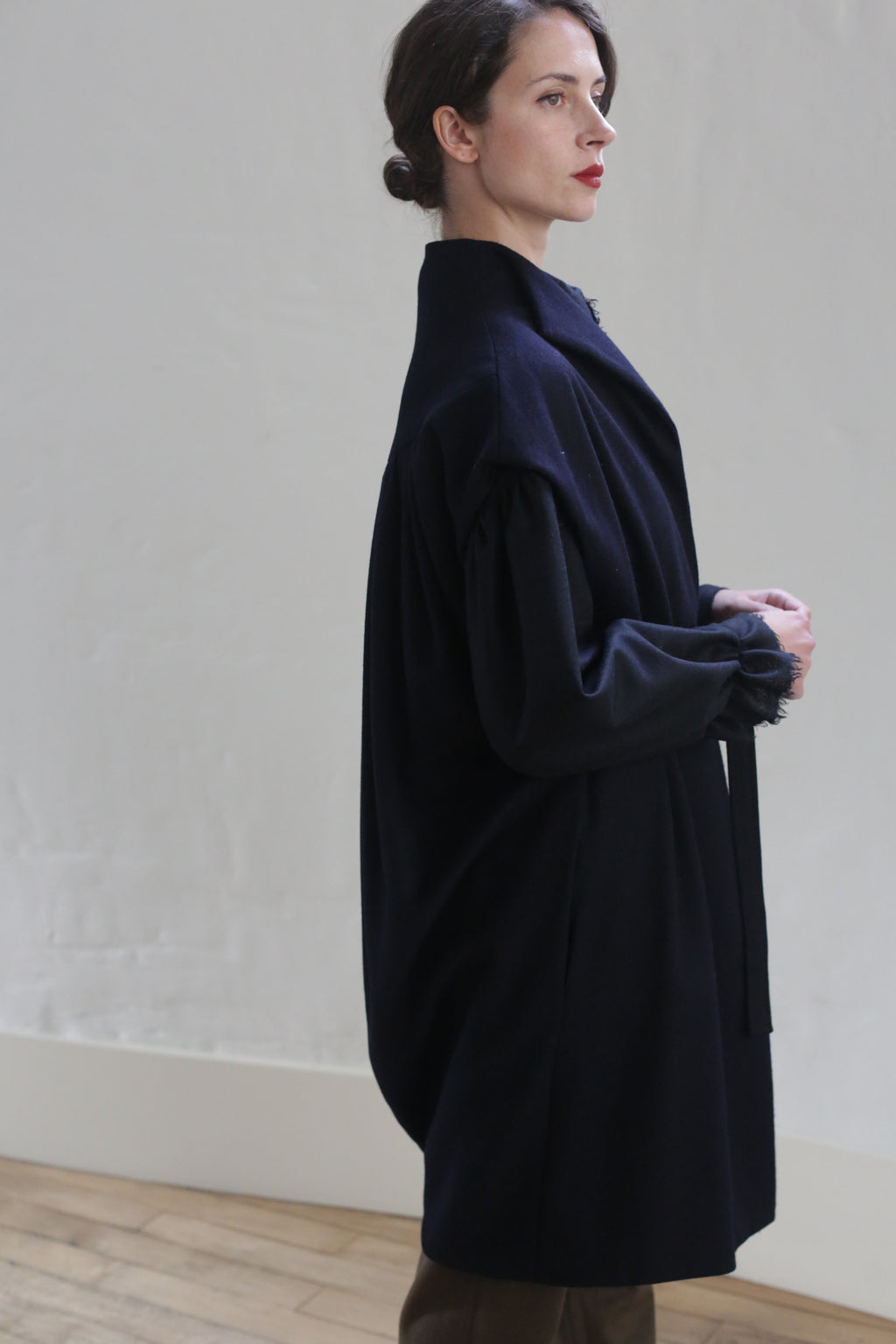 A woman with short, dark hair is standing indoors, facing left. She is wearing a long, dark Poet Vest Coat | Navy from Atelier Lan Jaenicke and has her hands clasped in front of her. The background features a plain, light-colored wall and a wooden floor.