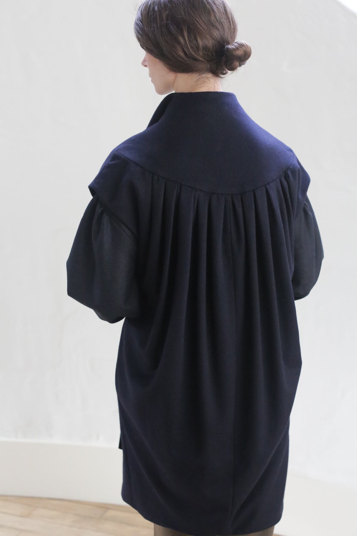 A person with dark hair styled in a low bun is standing with their back facing the camera. They are wearing a dark, loose-fitting garment with pleated details on the back, reminiscent of a Poet Vest Coat | Navy. The background appears to be a white wall with a wooden floor.