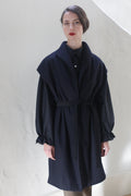 A woman with red lipstick and dark hair styled back is wearing a Poet Vest Coat | Navy with a tied waist and gathered sleeves. She stands against a light-colored wall, her expression appearing neutral.