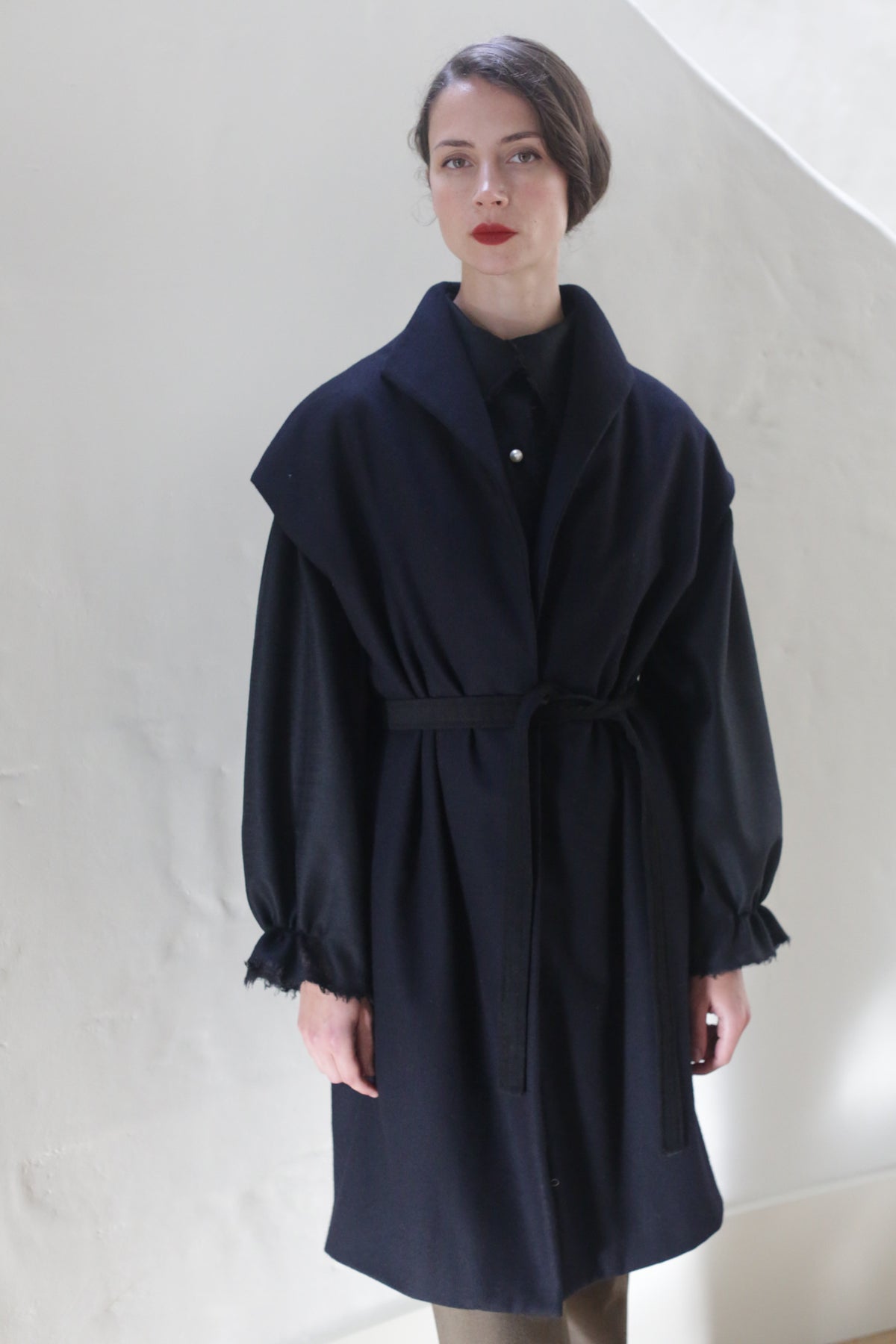 A woman with red lipstick and dark hair styled back is wearing a Poet Vest Coat | Navy with a tied waist and gathered sleeves. She stands against a light-colored wall, her expression appearing neutral.