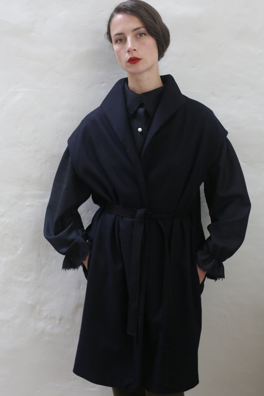 A person stands against a white wall, wearing an Atelier Lan Jaenicke Poet Vest Coat | Navy over a dark outfit with fringed sleeves. They have short, slicked-back hair and red lipstick, with hands in coat pockets, gazing at the camera.