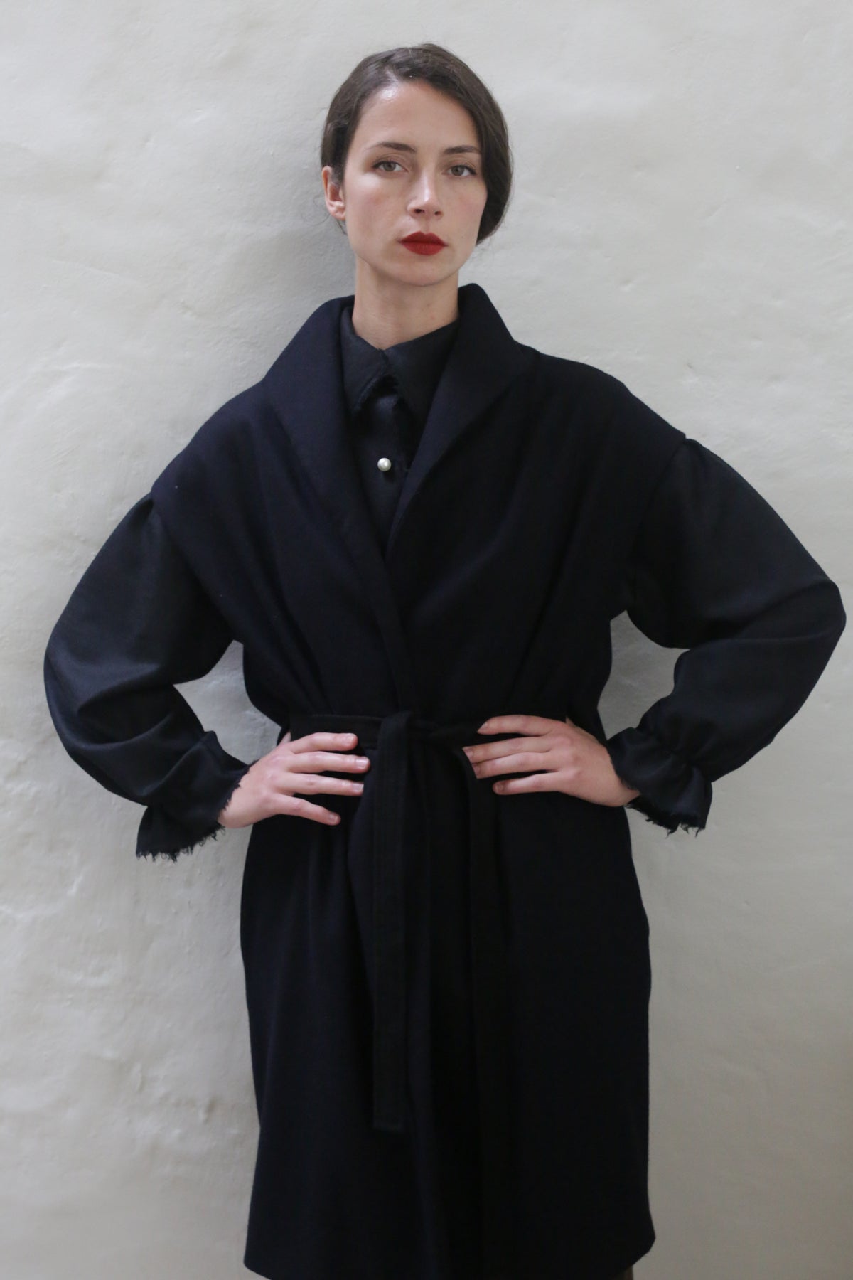 A person stands against a plain light-colored wall, wearing a Poet Vest Coat | Navy with a belt tied around the waist. Their hands are placed on their hips, and they have short dark hair and are wearing red lipstick. They exude confidence in the handmade cashmere coat.