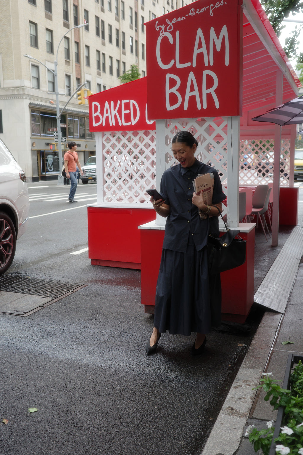 A woman stands on a city sidewalk by a red and white "Clam Bar" sign, looking at her phone with a newspaper in hand. She wears the NYFW Edition — Work Shirt & Wrap Skirt Set made from Japanese typewriter cotton and carries a black purse.