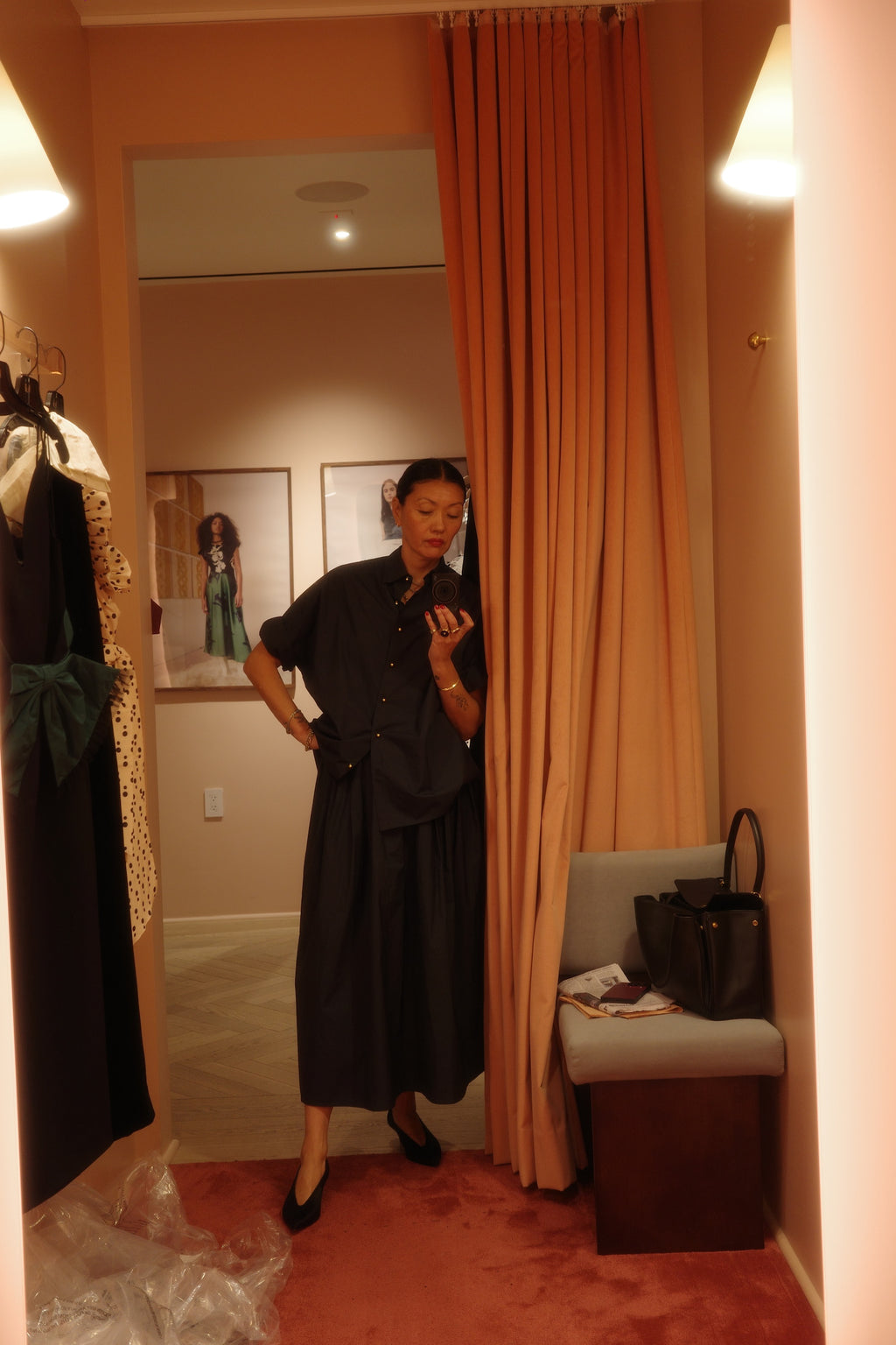 A woman wearing the NYFW Edition — Work Shirt & Wrap Skirt Set stands in a fitting room, looking at her phone. Clothes hang on the wall, a black handbag sits on the bench, and peach-hued curtains warm the scene.