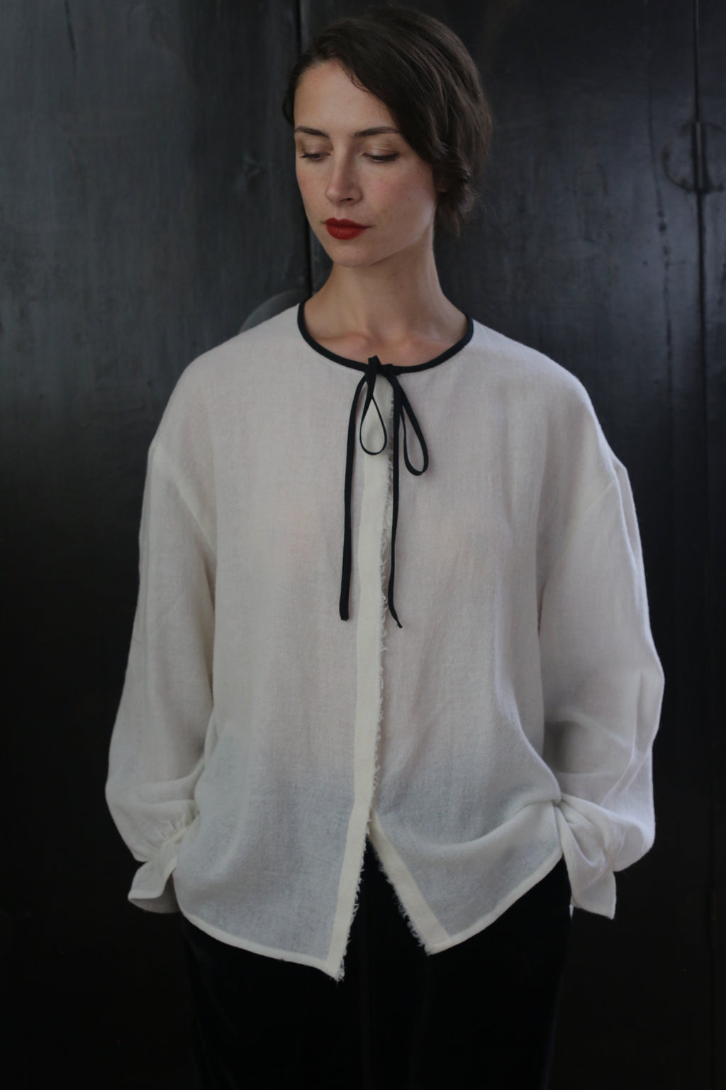 Person standing against a dark background, wearing a sheer, white long-sleeve blouse with a black neckline tie and black pants. The Ribbon Shirt - Japanese Wool Gauze seems inspired by traditional Ribbon Shirts. They have short, dark hair, and their hands are in their pockets. The person has a relaxed, contemplative expression with eyes cast downward.