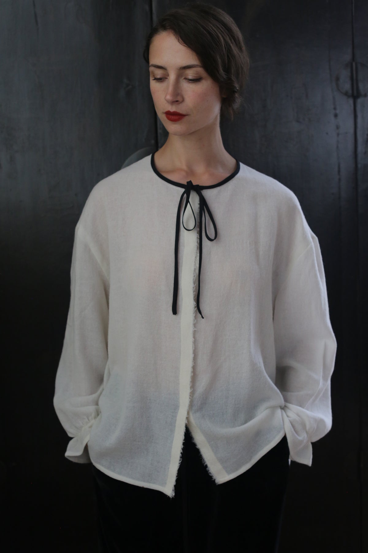 Person standing against a dark background, wearing a sheer, white long-sleeve blouse with a black neckline tie and black pants. The Ribbon Shirt - Japanese Wool Gauze seems inspired by traditional Ribbon Shirts. They have short, dark hair, and their hands are in their pockets. The person has a relaxed, contemplative expression with eyes cast downward.