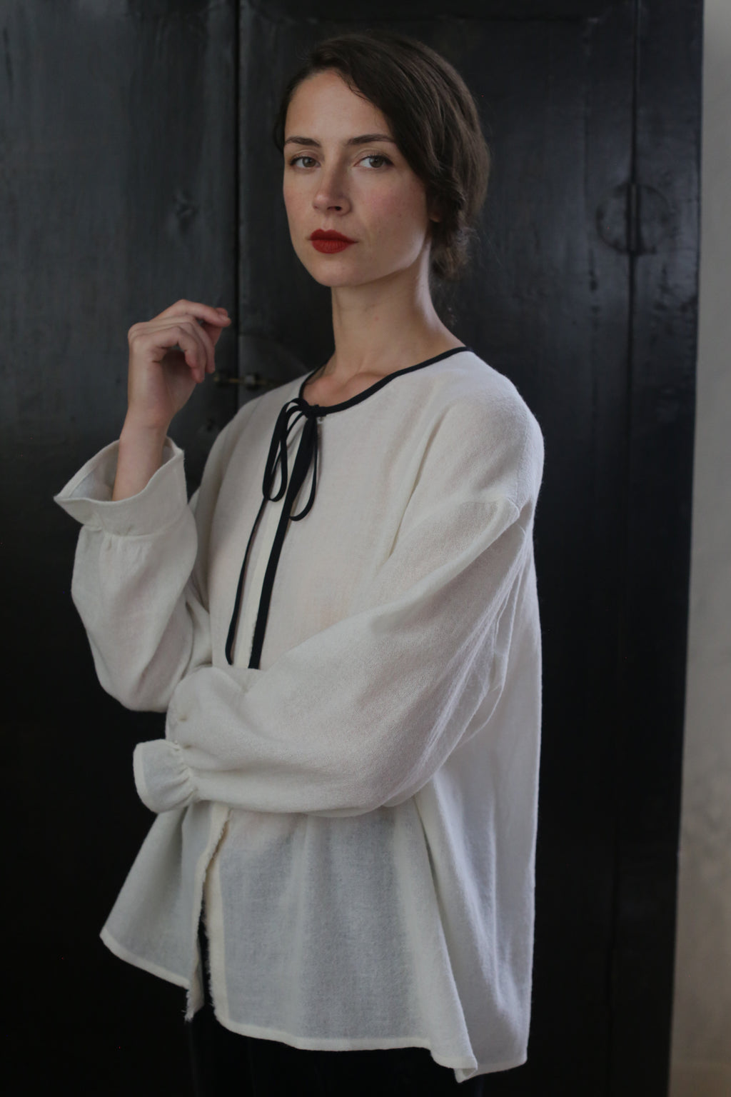 A woman with dark hair, styled in a loose updo, stands against a dark background. She wears a Ribbon Shirt - Japanese Wool Gauze with a black ribbon tie at the neckline. Her expression is serene, and she has red lipstick on. One hand is raised near her face.